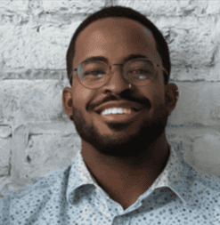 Smiling man with glasses in front of a white brick wall.