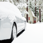 White car covered in snow in a winter landscape with trees and a house.