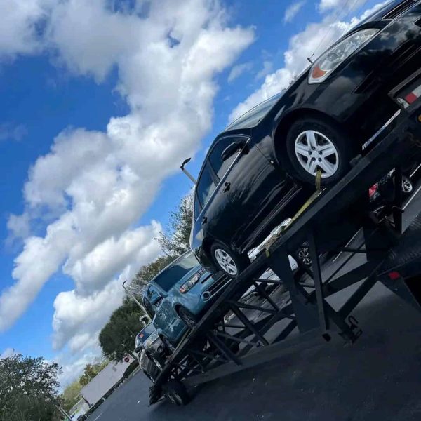 Black car on a car carrier trailer during daytime with blue sky and clouds.