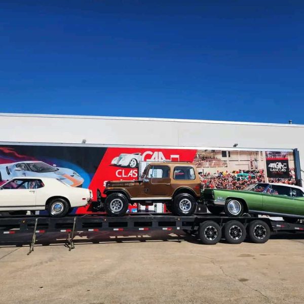 Classic cars loaded on a transport trailer outside a logistics facility.
