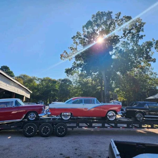 Vintage cars being transported on a flatbed trailer in a sunny outdoor setting.